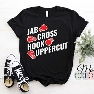 May include: Black t-shirt with the words "JAB CROSS HOOK UPPERCUT" in white, accompanied by red boxing glove graphics. A pair of black and white sneakers are in the foreground. The shirt also has the words "More Colors" in the bottom right corner.
