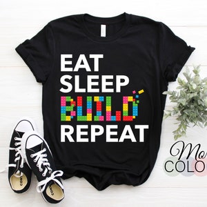 May include: Black t-shirt with white text that reads "Eat Sleep Build Repeat". The word "Build" is made up of colourful toy building blocks.