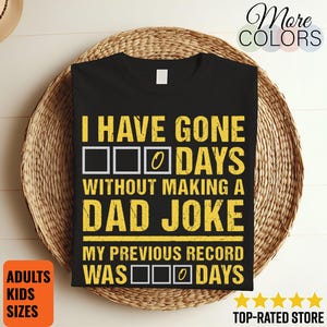 May include: Black t-shirt with yellow text that reads "I HAVE GONE 0 DAYS WITHOUT MAKING A DAD JOKE." The text "MY PREVIOUS RECORD WAS 0 DAYS" is also displayed. The shirt is on a woven basket.