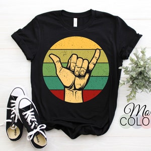 May include: Black t-shirt with a vintage-style graphic of a hand making the "hang loose" gesture in front of a sunset. The graphic is in shades of yellow, green, red, and brown.