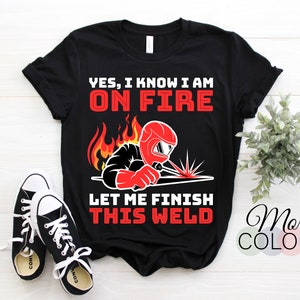 May include: Black t-shirt with a red and white graphic of a welder with flames and the text "YES, I KNOW I AM ON FIRE LET ME FINISH THIS WELD".