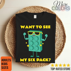 May include: Black t-shirt with a cartoon block design. The block is teal with sunglasses and flexed arms. Text reads "WANT TO SEE MY SIX PACK?" with "ADULTS KIDS SIZES" in an orange box. The words "More COLORS" are in the top right.