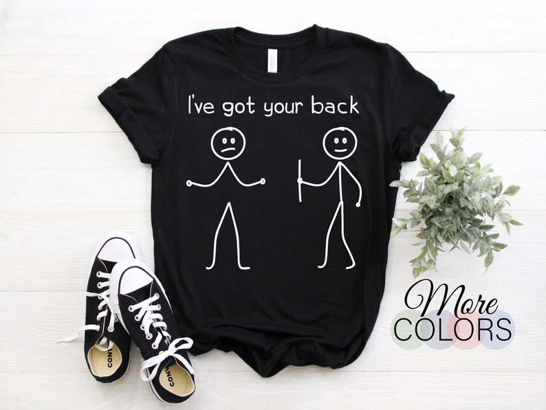 May include: Black t-shirt with white text that reads "I've got your back" and a stick figure illustration. The shirt is lying flat on a white wooden surface with a pair of black trainers and a small green plant.