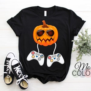 May include: Black t-shirt featuring a cartoon pumpkin wearing sunglasses and holding two white video game controllers. The pumpkin has an orange face with a carved-out mouth and is a Halloween-themed design. The text "More Colors" is on the right.
