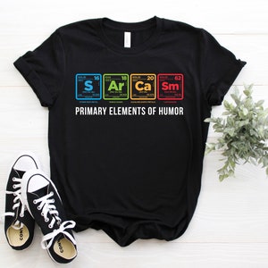 Puede incluir: Camiseta negra con un diseño de temática científica que presenta los elementos químicos "S", "Ar", "Ca" y "Sm" dispuestos en un formato de tabla periódica. El texto "PRIMARY ELEMENTS OF HUMOR" está impreso debajo de los elementos.