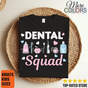 Puede incluir: Camiseta negra con las palabras "DENTAL Squad" en blanco y rosa, con dibujos de herramientas dentales y corazones. La camiseta se muestra sobre una cesta tejida. Las palabras "ADULTOS TALLAS NIÑOS" están en un recuadro naranja.