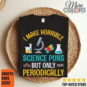 May include: Black t-shirt with the text "I make horrible science puns but only periodically" in yellow and blue. The shirt features science-themed graphics including a microscope, beakers, and molecular models. Available in adult and kids sizes.