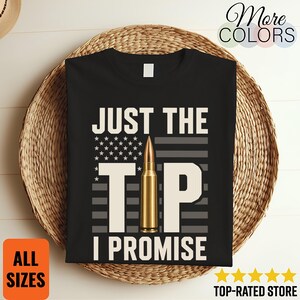 May include: A black t-shirt featuring a graphic design with the text JUST THE TIP I PROMISE in white capital letters. The letter I is replaced by a gold-coloured bullet. Behind the text is a stylised American flag in shades of grey and white.