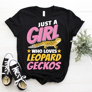 Puede incluir: Camiseta negra con texto rosa que dice "Just a Girl Who Loves Leopard Geckos". Se muestra un gecko leopardo en la camiseta.