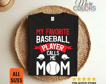May include: Black t-shirt with the text "MY FAVORITE BASEBALL PLAYER CALLS ME MOM" in white and red. The design includes a baseball, baseball caps, and the word "MOM" in large white letters. The shirt is displayed on a woven basket.