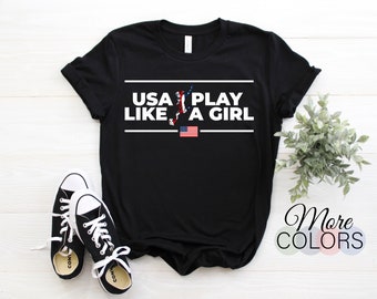 May include: Black t-shirt with white text that reads "USA Play Like A Girl" with a silhouette of a soccer player and an American flag.