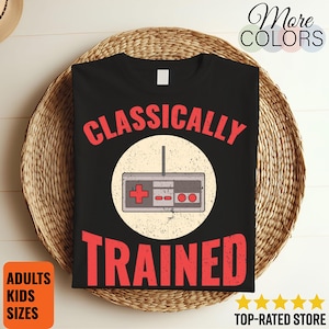 May include: Black t-shirt with the words "CLASSICALLY TRAINED" in red, surrounding a retro video game controller graphic. The shirt is displayed on a woven basket. Additional text includes "ADULTS KIDS SIZES" and "TOP-RATED STORE".