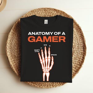 K&ouml;nnte beinhalten: Schwarzes T-Shirt mit dem Text "Anatomy of a Gamer" in Wei&szlig; und einer Skeletthand mit den Tasten "W, S, A, D, Shift, Ctrl, Space" in Orange.