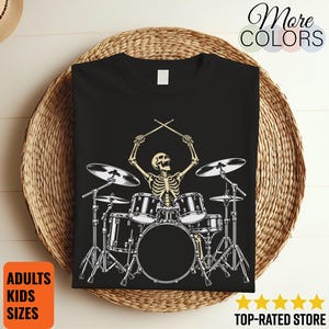 Può includere: Una t-shirt nera con una stampa grafica di uno scheletro che suona la batteria. Lo scheletro è posizionato al centro, tenendo le bacchette in aria sopra i tamburi. Il set di batteria include una grancassa, rullante, tom e piatti.
