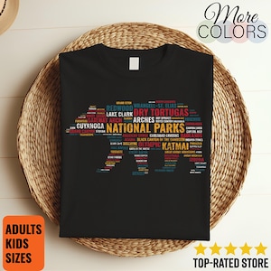 Könnte beinhalten: Schwarzes T-Shirt mit einem farbenfrohen Grafikdesign der US-Nationalparks, darunter Redwood, Arches und Olympic. Der Text "NATIONAL PARKS" ist prominent. Zusätzlicher Text enthält "ADULTS KIDS SIZES" und "TOP-RATED STORE".