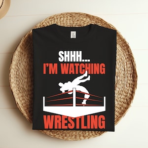 Op de afbeelding: Zwart T-shirt met witte tekst die "SHHH... I'M WATCHING WRESTLING" luidt. Boven de tekst bevindt zich een wit silhouet van twee worstelaars in een worstelring.