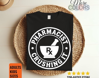 May include: A black t-shirt featuring a white circular graphic with the text PHARMACIST CRUSHING IT surrounding a mortar and pestle icon with the Rx symbol centred inside. The design is printed in a bold, clean font on the front of the garment.