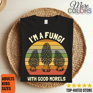 May include: Black t-shirt with a graphic featuring three morel mushrooms set against a sunset design. The text reads "I'M A FUNGI WITH GOOD MORELS". Additional text indicates adult and kids sizes are available.