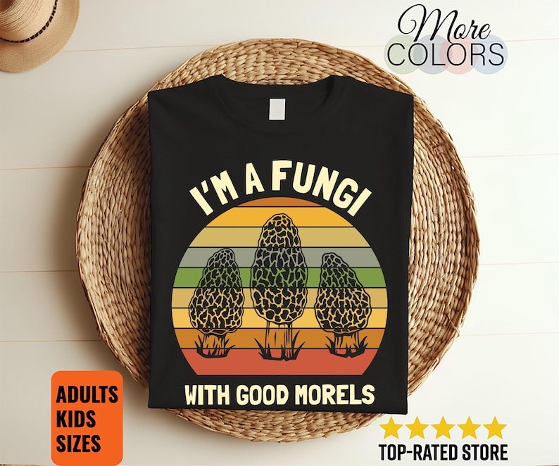 Puede incluir: Camiseta negra con un dise&ntilde;o de tres setas colmenilla sobre un fondo de puesta de sol. El texto dice "I'M A FUNGI WITH GOOD MORELS". Disponible en tallas para adultos y ni&ntilde;os.