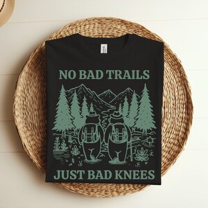 May include: Black t-shirt featuring a green graphic of two bears hiking in a forest. The text "NO BAD TRAILS JUST BAD KNEES" is printed on the shirt.