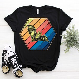 May include: Black t-shirt featuring a retro design of a bat inside a hexagon with colorful stripes. The bat is yellow and the stripes are orange, red, and blue. A pair of black and white sneakers are in the foreground.