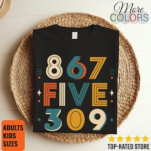 Op de afbeelding: Een zwart t-shirt met een grafisch ontwerp met de tekst 867 FIVE 309 in een retro-geïnspireerd lettertype. De cijfers en letters zijn gekleurd in tinten mosterdgeel, blauwgroen en oranje, geaccentueerd door kleine ster- en lijnsymbolen.