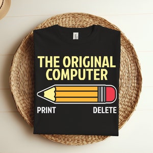 May include: Black t-shirt with the text "THE ORIGINAL COMPUTER" in yellow letters. A yellow pencil with a red eraser is below the text. The words "PRINT" and "DELETE" are below the pencil.