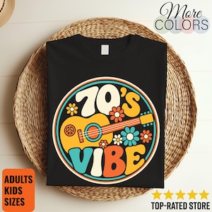 May include: A black t-shirt featuring a circular graphic with the text 70's VIBE in a bold, rounded font. The design includes a yellow acoustic guitar, a peace sign, and various colourful flowers in shades of orange, blue, and cream, all enclosed within a multi-coloured circular border.