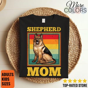 May include: Black t-shirt featuring a German Shepherd dog illustration within a retro-style frame with a red, orange, and yellow striped background. The text "SHEPHERD MOM" is displayed in yellow. The shirt also has the text "ADULTS KIDS SIZES".