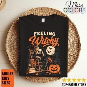 May include: A black t-shirt featuring the text Feeling Witchy in orange and white. The graphic depicts a person in a witch hat sitting in a rocking chair, drinking from a mug, with a black cat, a lantern, books, a pumpkin, and a full moon.