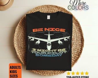 May include: Black t-shirt with a graphic of a white airplane and the text "BE NICE I MIGHT BE YOUR PILOT SOMEDAY". The shirt also has the words "ADULTS KIDS SIZES" in an orange box. The words "More COLORS" are in the top right corner.