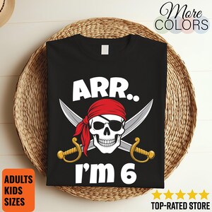 May include: A black t-shirt featuring a white graphic of a skull wearing a red bandana and an eye patch, positioned in front of two crossed silver swords with gold hilts. The text ARR.. I'M 6 is printed in white above and below the graphic.