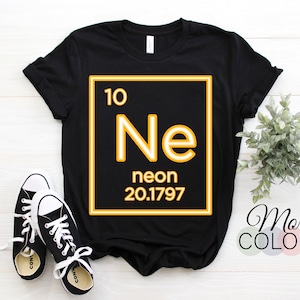 May include: Black t-shirt featuring a yellow periodic table element graphic. The element is neon (Ne) with the atomic number 10 and atomic weight 20.1797. The text "More Colors" is visible in the bottom right corner of the image.