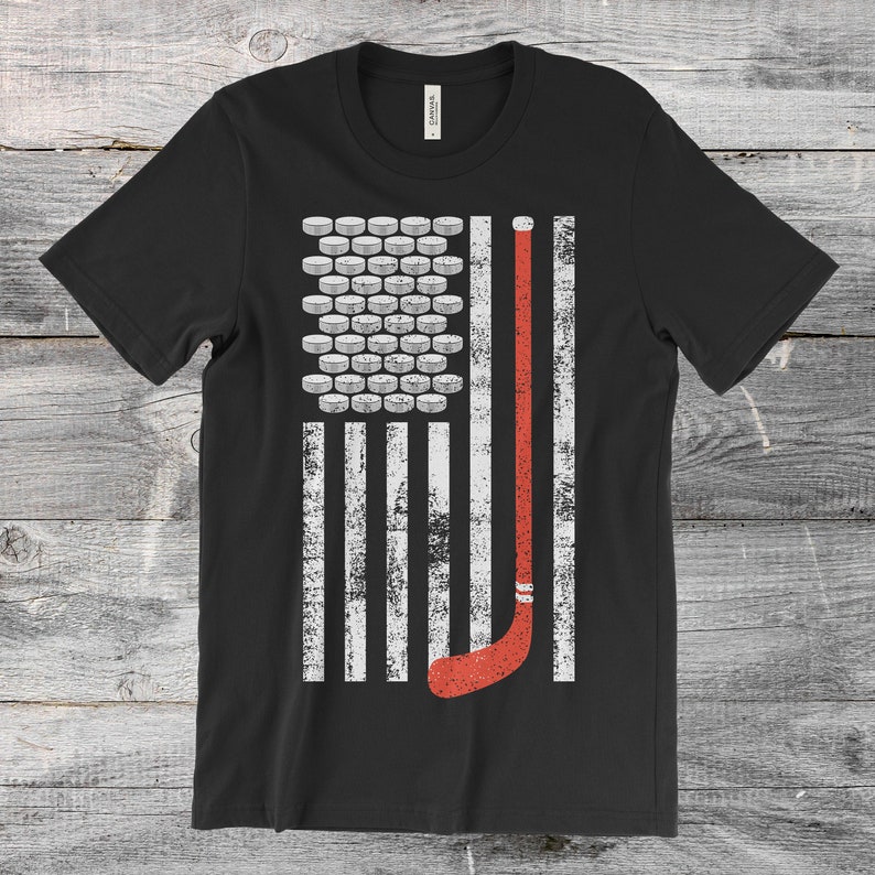 May include: Black t-shirt featuring a distressed graphic of the American flag, with hockey pucks replacing the stars and a hockey stick in place of a stripe. The design is in white and red.