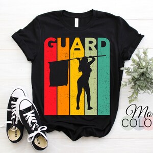 May include: Black t-shirt with a retro design featuring the word "GUARD" above a silhouette of a person holding a flag. The design incorporates vertical stripes in red, orange, yellow, and teal. A pair of black and white sneakers are in the foreground.