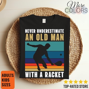 Puede incluir: Camiseta negra con el texto "NEVER UNDERESTIMATE AN OLD MAN WITH A RACKET" y una silueta de un jugador de tenis. El diseño utiliza una combinación de colores retro con rayas azules, turquesas, naranjas y rojas. La camiseta se muestra sobre una cesta tejida.