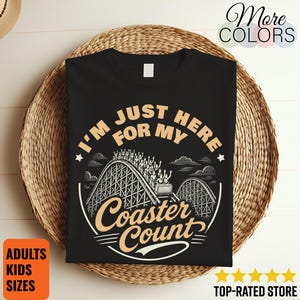 Puede incluir: Una camiseta negra con un estampado gráfico en color beige claro. El diseño muestra una montaña rusa con pasajeros y el texto "I'M JUST HERE FOR MY Coaster Count" dispuesto en un diseño circular con estrellas y nubes.