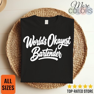Könnte beinhalten: Ein schwarzes kurzärmeliges T-Shirt mit dem weißen Schriftzug World's Okayest Bartender in einer stilisierten Schreibschrift mittig auf der Brust. Das Kleidungsstück hat einen einfachen Rundhalsausschnitt und einen geraden Saum.