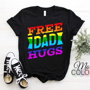 Könnte beinhalten: Schwarzes T-Shirt mit dem Text "FREE DAD HUGS" in Regenbogenfarben. Der Text ist in einer fetten, blockartigen Schrift.