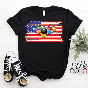 May include: Black t-shirt featuring a distressed American flag design with billiard balls and cues. The design incorporates the number 8 ball prominently. A pair of black and white sneakers are in the foreground. The words "More Colors" are in the bottom right corner.