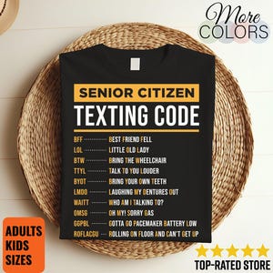 May include: Black t-shirt with the text "SENIOR CITIZEN TEXTING CODE" in white and yellow. The shirt lists texting acronyms and their meanings, such as "BFF - BEST FRIEND FELL" and "LOL - LITTLE OLD LADY". The shirt is displayed on a woven basket.