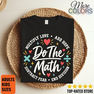 May include: A black t-shirt featuring a graphic design with the text "Do The Math" in white, surrounded by the phrases "Multiply Love," "Add Hope," "Subtract Fear," and "End Division" in white, accented with red hearts and colourful math symbols.