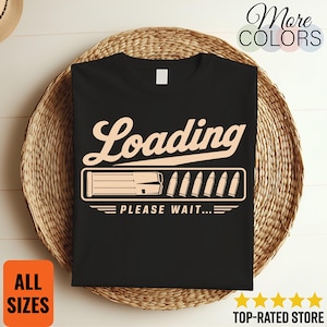 Peut inclure: Un t-shirt noir avec un motif graphique beige clair représentant le mot Loading en écriture cursive au-dessus d'une barre de chargement. La barre contient un chargeur et plusieurs balles, suivis du texte PLEASE WAIT en majuscules.