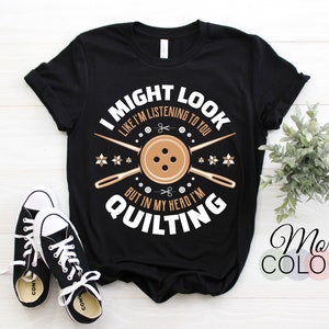 Puede incluir: Camiseta negra con el texto blanco "I MIGHT LOOK LIKE I'M LISTENING TO YOU BUT IN MY HEAD I'M QUILTING". El diseño incluye un botón, agujas y tijeras. Un par de zapatillas negras y blancas están en primer plano.