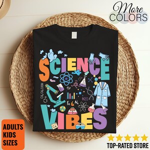 Pode incluir: Camiseta preta com as palavras "SCIENCE VIBES" em letras coloridas. O design inclui ilustrações de temática científica, como um microscópio e símbolos atómicos. A camiseta é exibida em uma cesta de vime.
