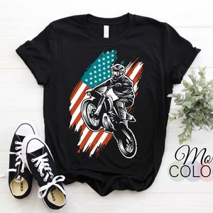 May include: Black t-shirt featuring a motocross rider graphic with an American flag design. The graphic is in white and the flag has red and blue accents. The shirt is paired with black and white sneakers.