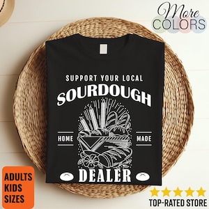 May include: Black t-shirt with white text and graphic. The text reads "SUPPORT YOUR LOCAL SOURDOUGH DEALER." The graphic depicts various types of bread. The shirt is displayed on a woven basket. The shirt is available in adult and kids sizes.