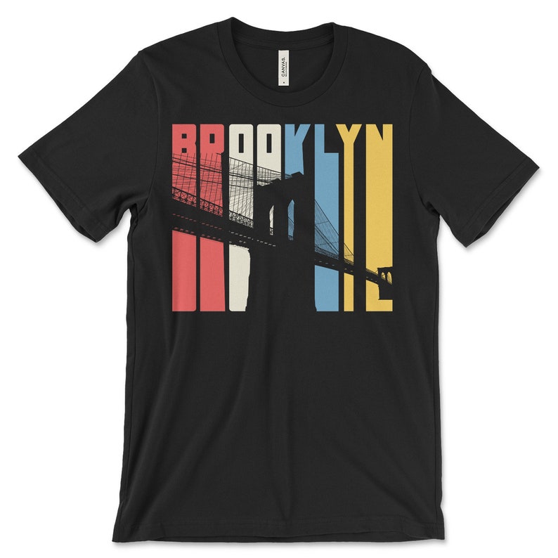 Op de afbeelding: Zwart T-shirt met een retro-grafische afbeelding van de Brooklyn Bridge in silhouet met het woord "BROOKLYN" in kleurrijke strepen.