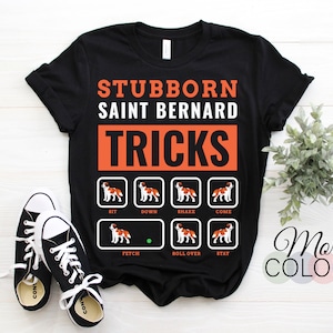 Könnte beinhalten: Schwarzes T-Shirt mit dem Text "STUBBORN SAINT BERNARD TRICKS" in Weiß und Orange. Das Shirt zeigt Illustrationen eines Bernhardiner-Hundes, der Tricks ausführt. Schwarze und weiße Turnschuhe sind im Vordergrund.