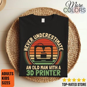 Op de afbeelding: Zwart T-shirt met een retro design met een 3D-printer en de tekst "NEVER UNDERESTIMATE AN OLD MAN WITH A 3D PRINTER". Het shirt wordt gepresenteerd op een gevlochten mand. Extra tekst bevat "ADULTS KIDS SIZES" en "TOP-RATED STORE".